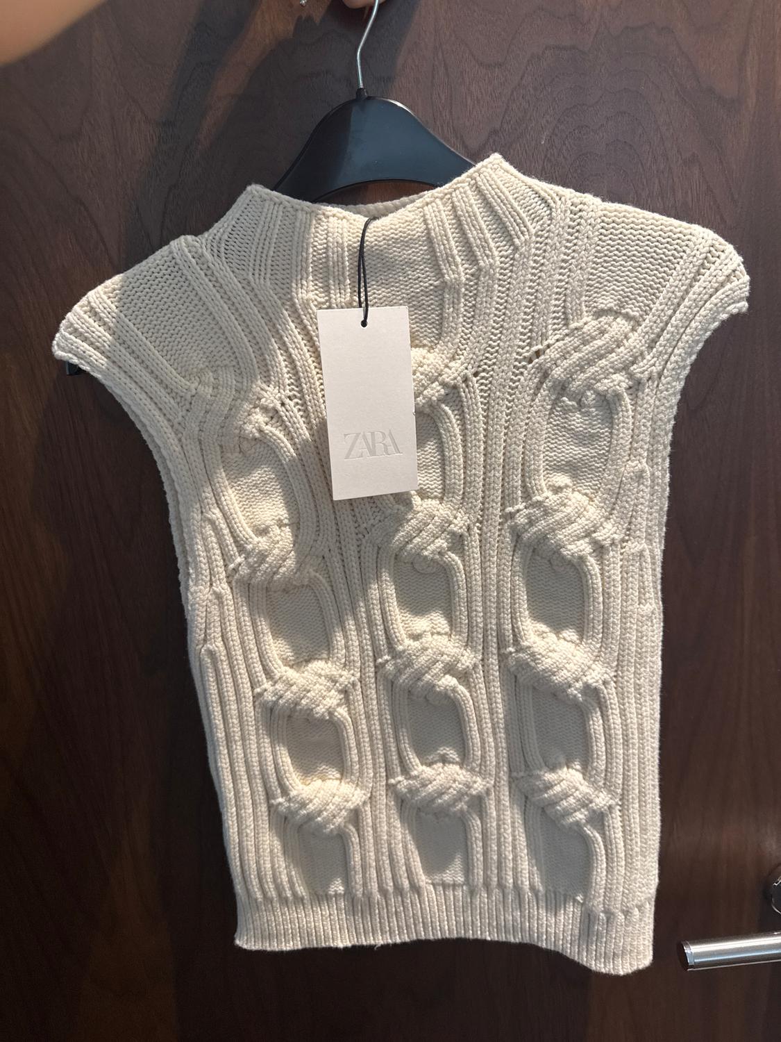Beige knitted cable-knit vest hanging on a black hanger. "ZARA" logo label visible. Wooden door background.