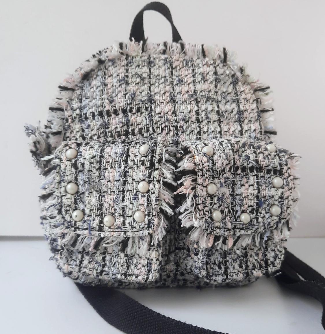 Textured fabric backpack in grey and pink tones, with fringes and front pockets decorated with pearls. White background.