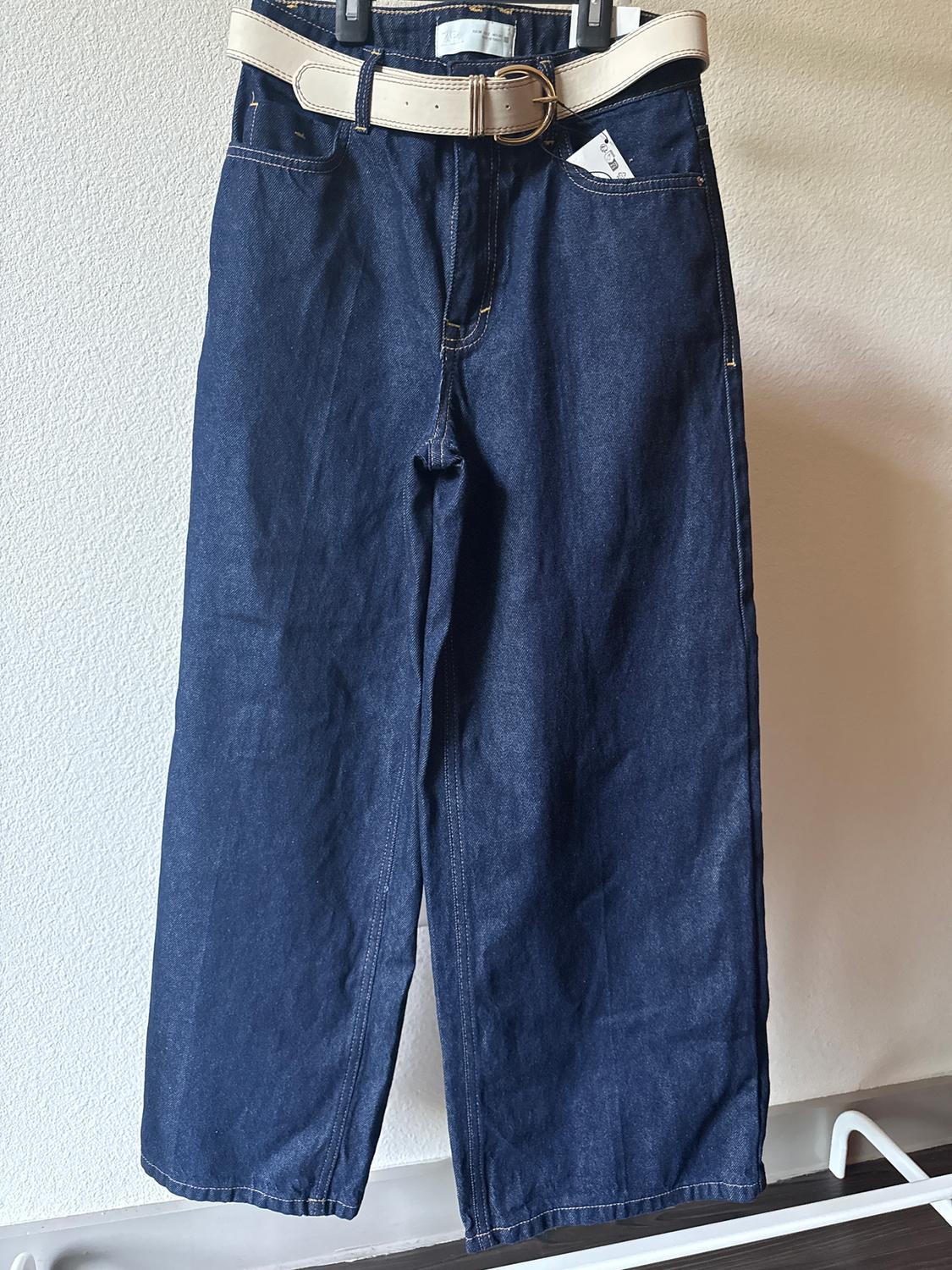 Dark blue jeans hanging on a hanger, with a beige belt and a visible label on the front pocket.