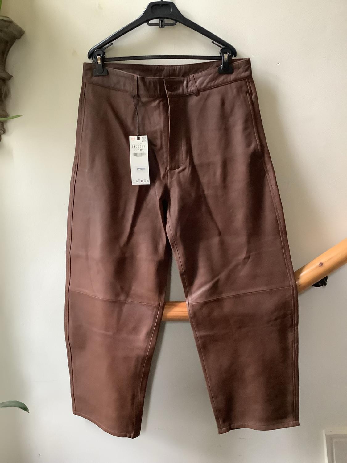 Brown trousers with a label hanging on a black hanger against a white wall, resting on a wooden railing.