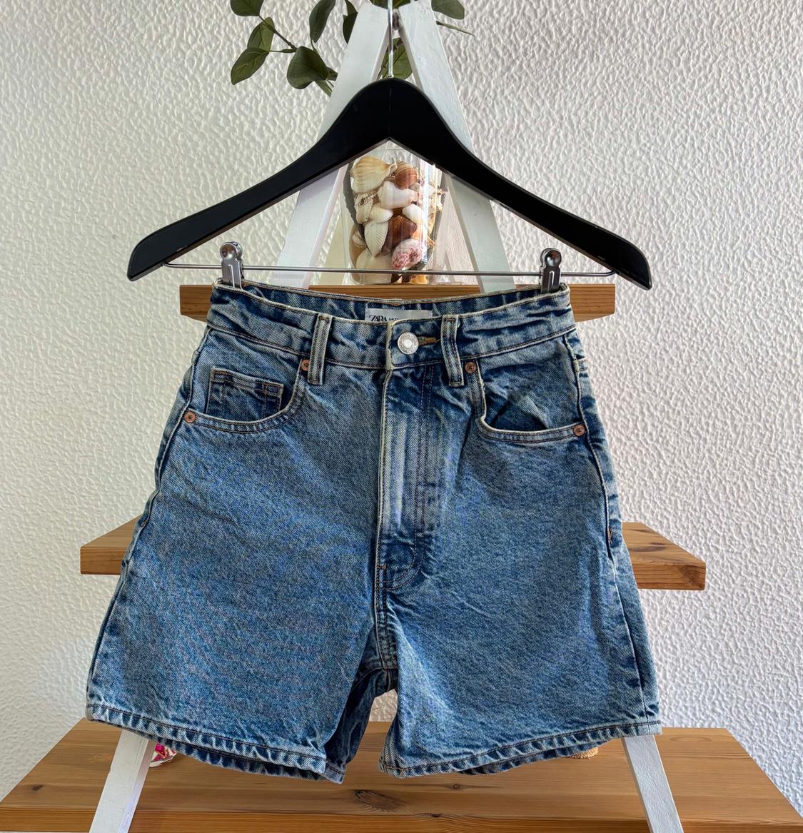Blue denim shorts hanging on a black hanger against a textured white wall background, with shells on display.