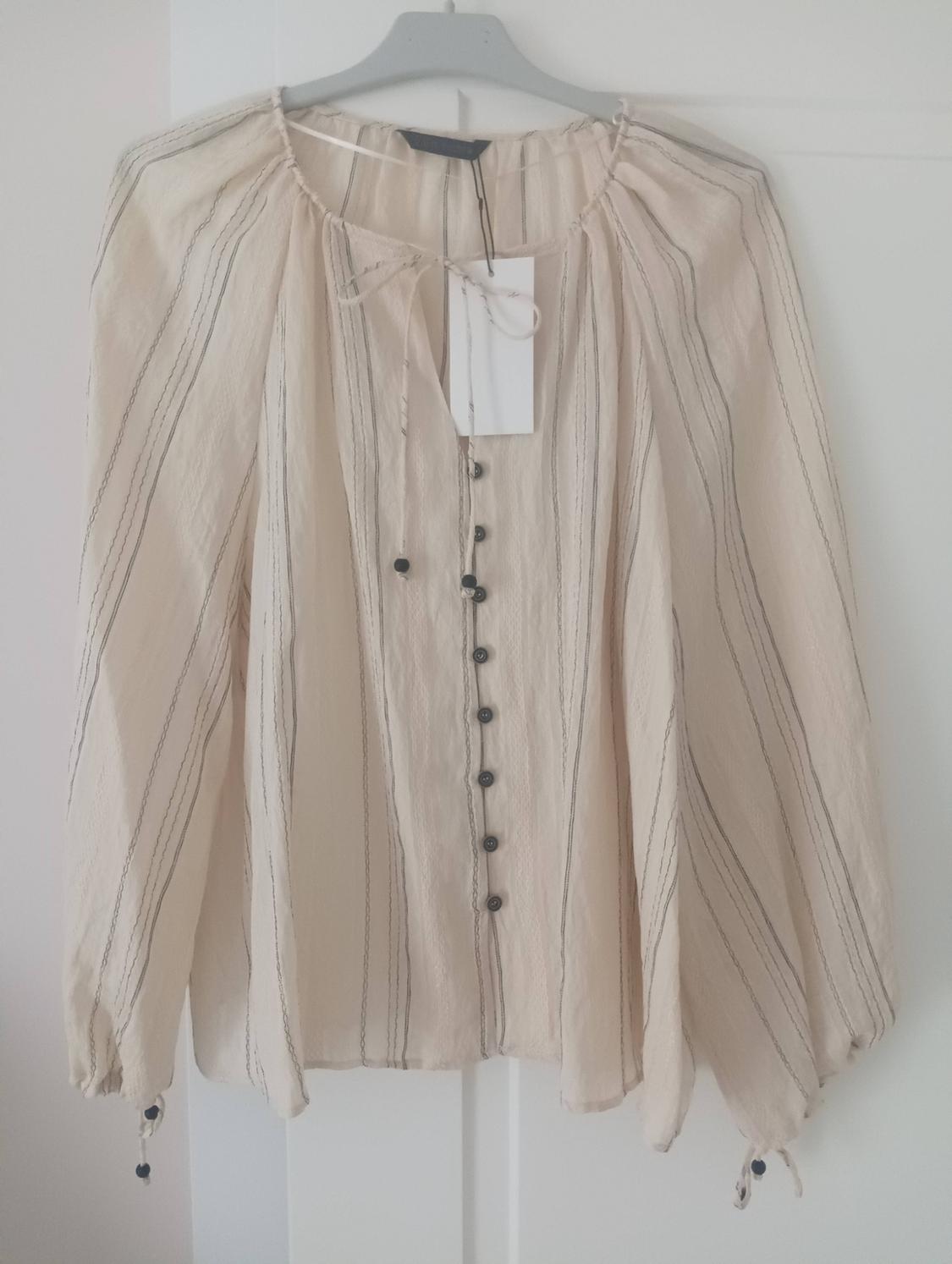Beige long-sleeved blouse with fine dark stripes, front buttons and ties at the neck and cuffs, hanging on a hanger against a white background.
