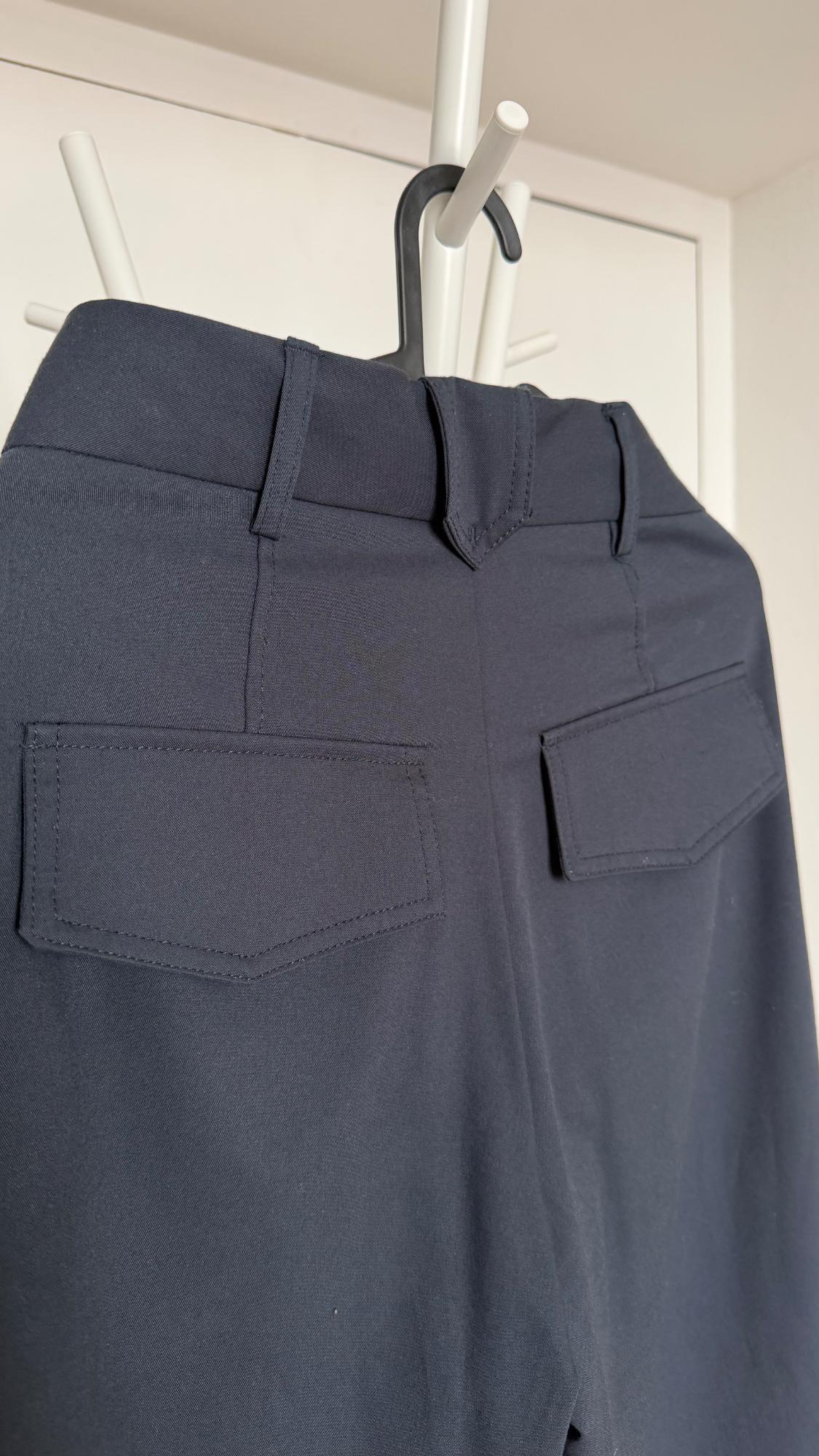 Rear view of navy blue trousers hanging on a white coat rack. The belt loops, V-detail, and flap pockets stand out.