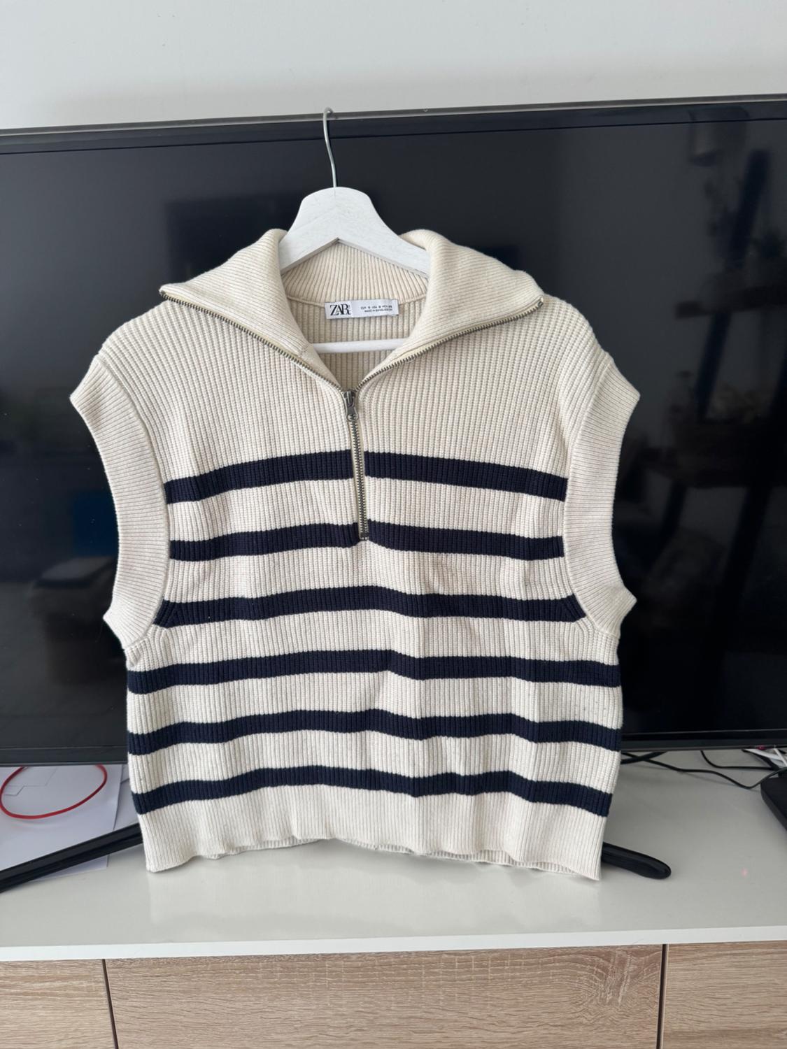 White knitted vest with dark stripes and a half-zip collar, hanging on a hanger in front of a television. ZARA text.