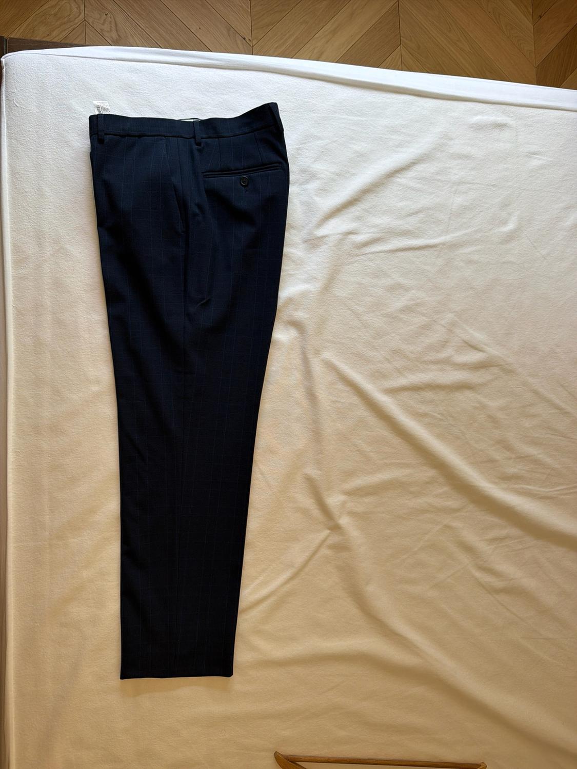 Dark trousers with subtle stripes folded over crumpled white fabric. A herringbone wooden floor and a hanger are visible.
