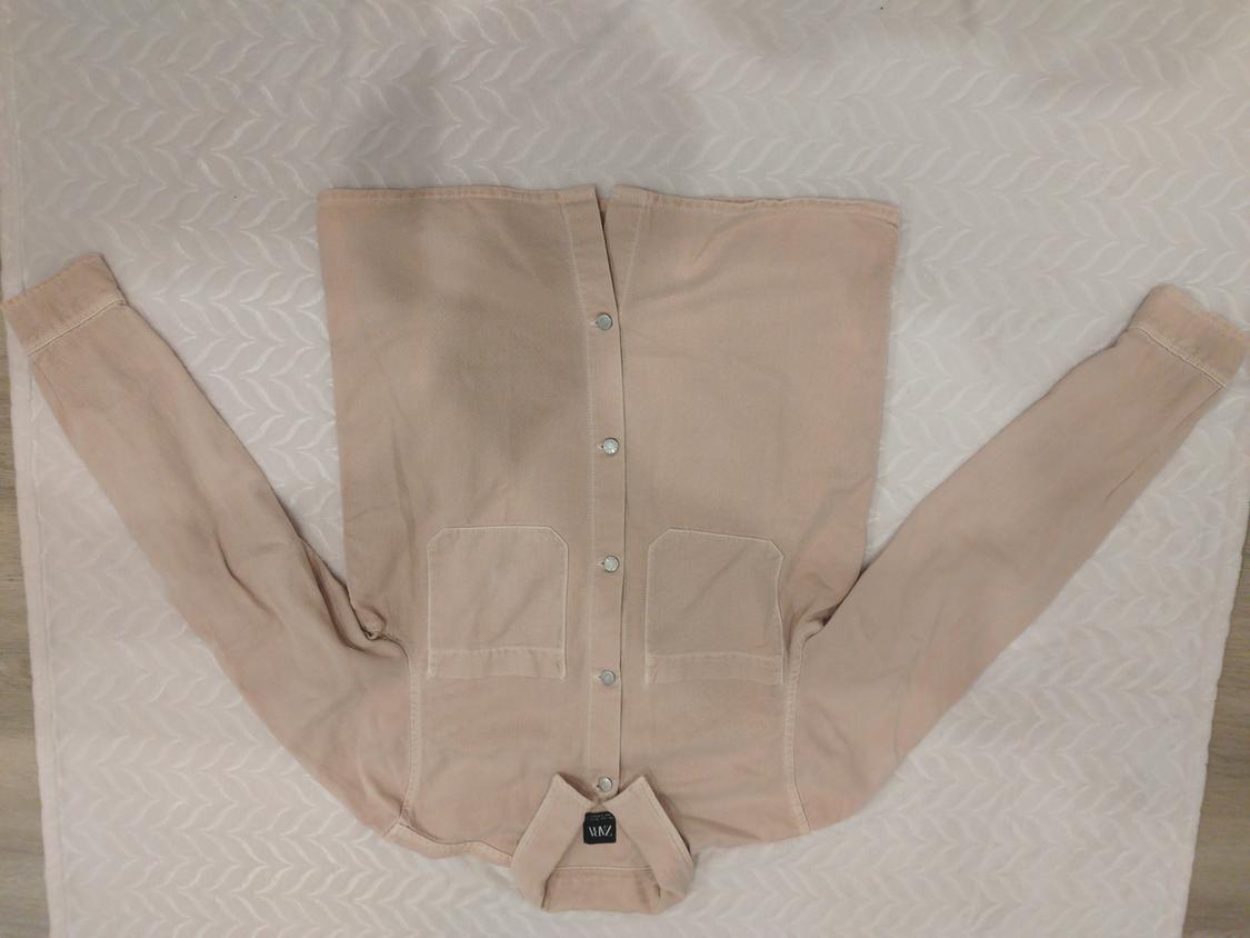 Beige long-sleeved shirt with buttons and two front pockets, placed on a surface with a leaf pattern. Label visible.