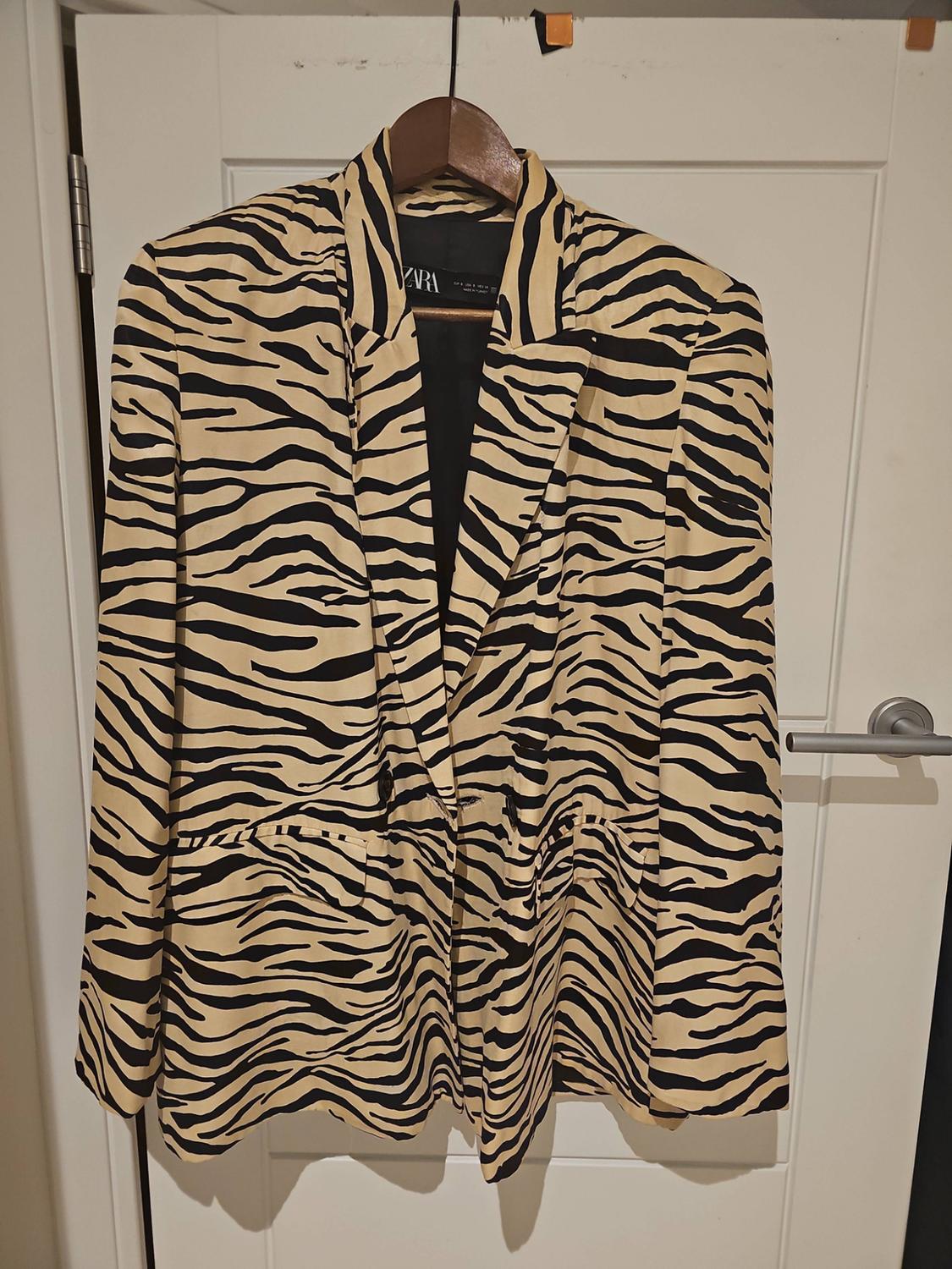 Beige and black zebra-print blazer hanging on a hanger over a white door. The inner label reads ZARA.