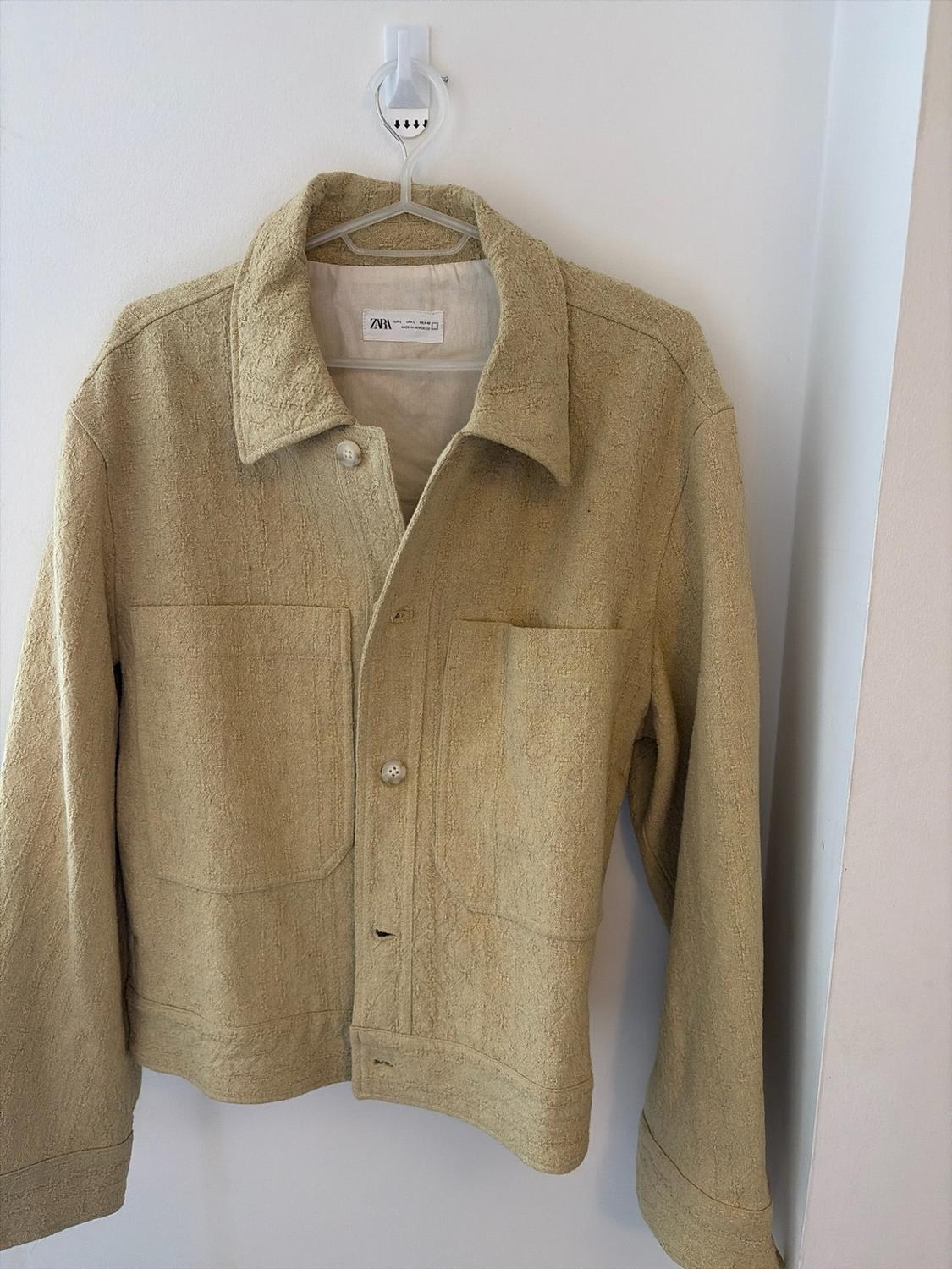 Textured beige jacket with buttons and front pockets, hanging on a hook against a white wall. Label with text ZARA.