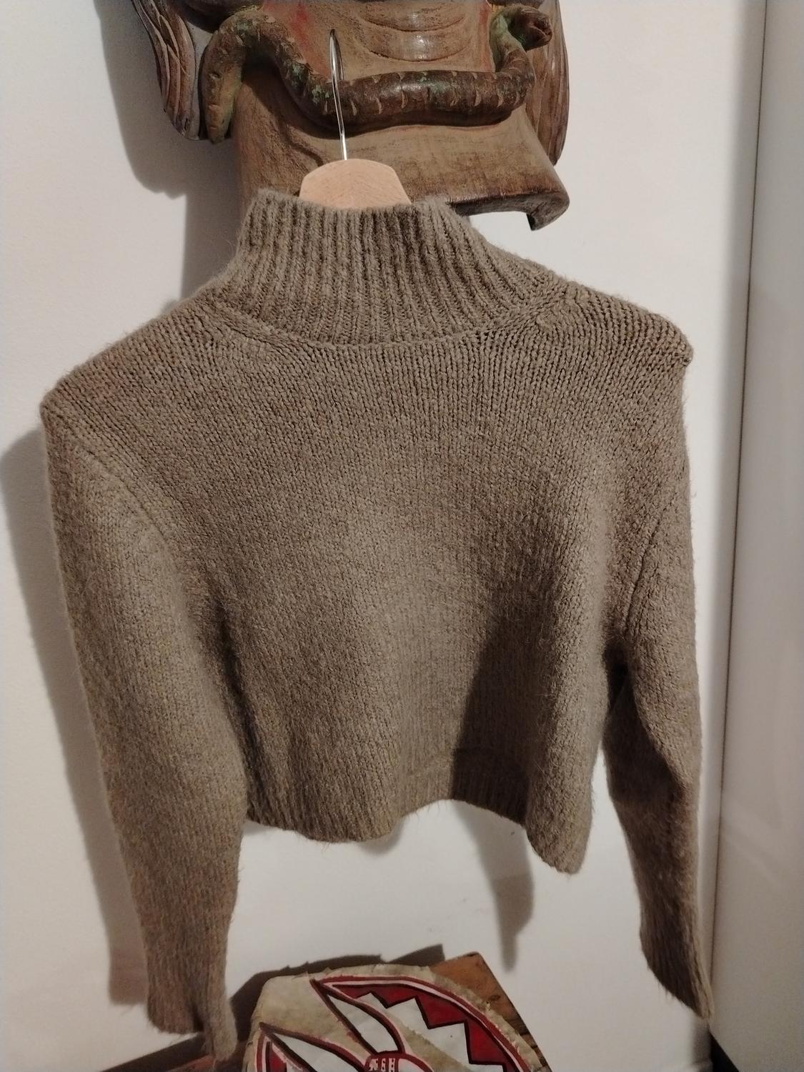 Brown knitted jumper hanging on a wooden hanger, with a high neck, against a white wall background and wooden decor.