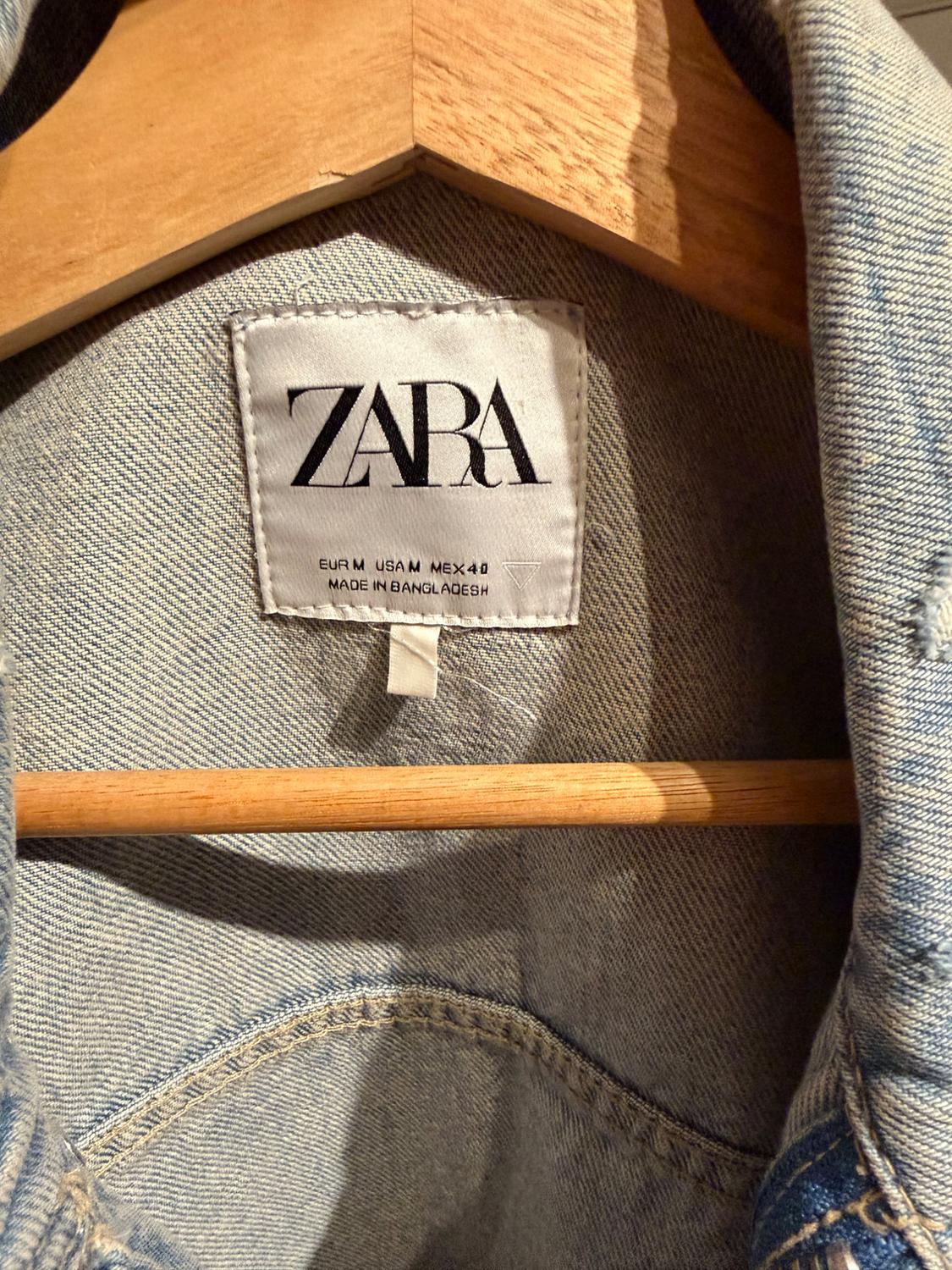 Denim jacket label on a wooden hanger, with text "ZARA", "EUR M USA M MEX 40", "MADE IN BANGLADESH".