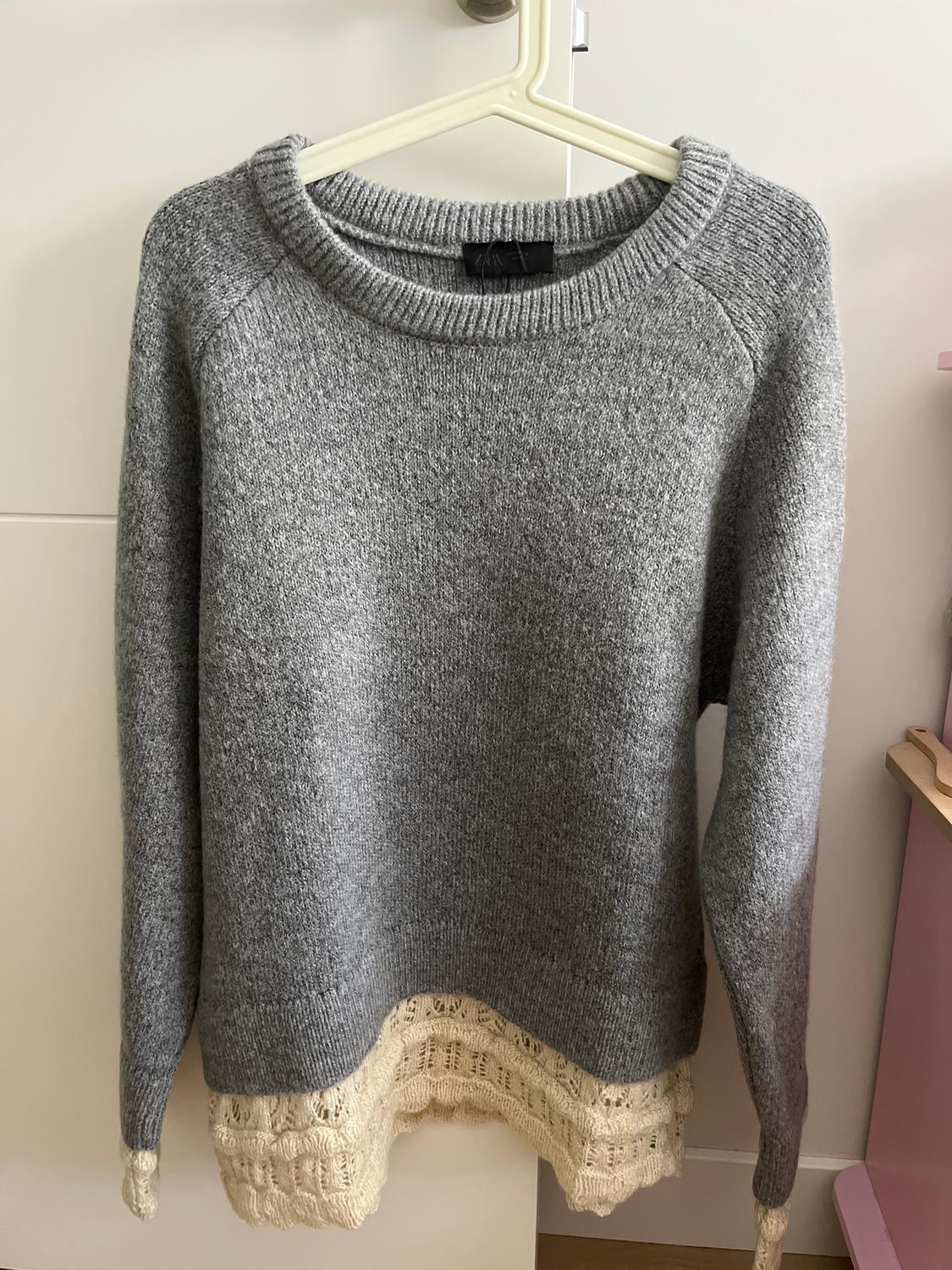 Chunky knit grey jumper with a round neck and beige lace detail on the hem, hanging on a white hanger.