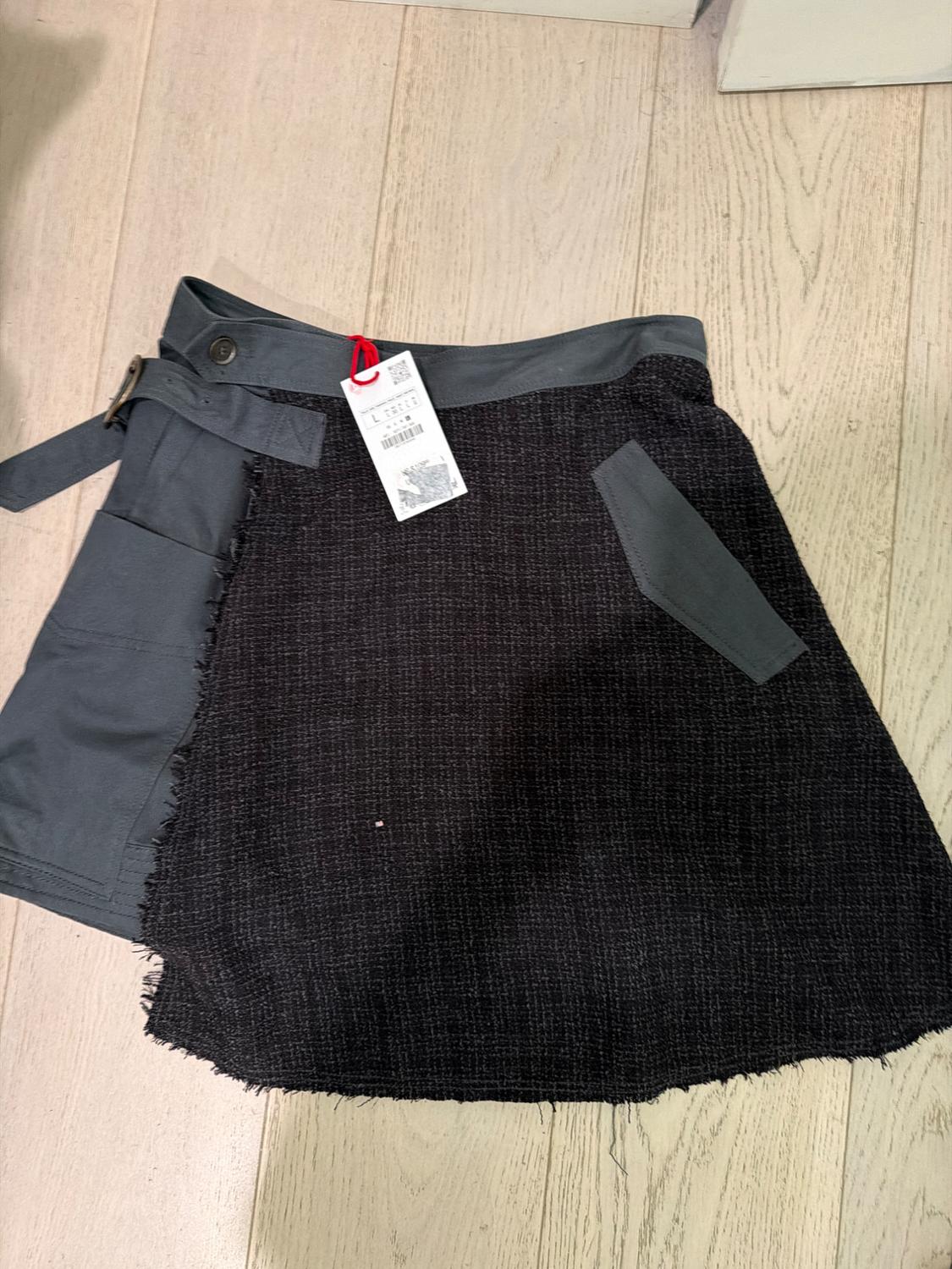 Short skirt on a wooden floor. Asymmetric design with a plain grey panel, buckle, textured dark fabric, frayed edges and label.