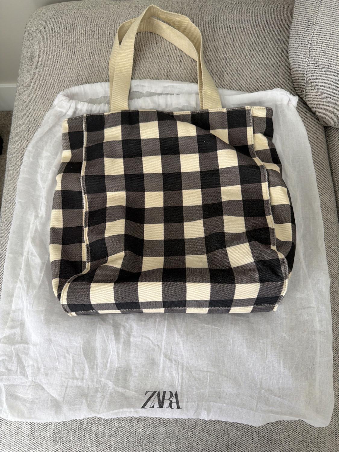 Black and white checked tote bag with light handles, on a white fabric dust bag with the text ZARA, resting on a grey sofa.