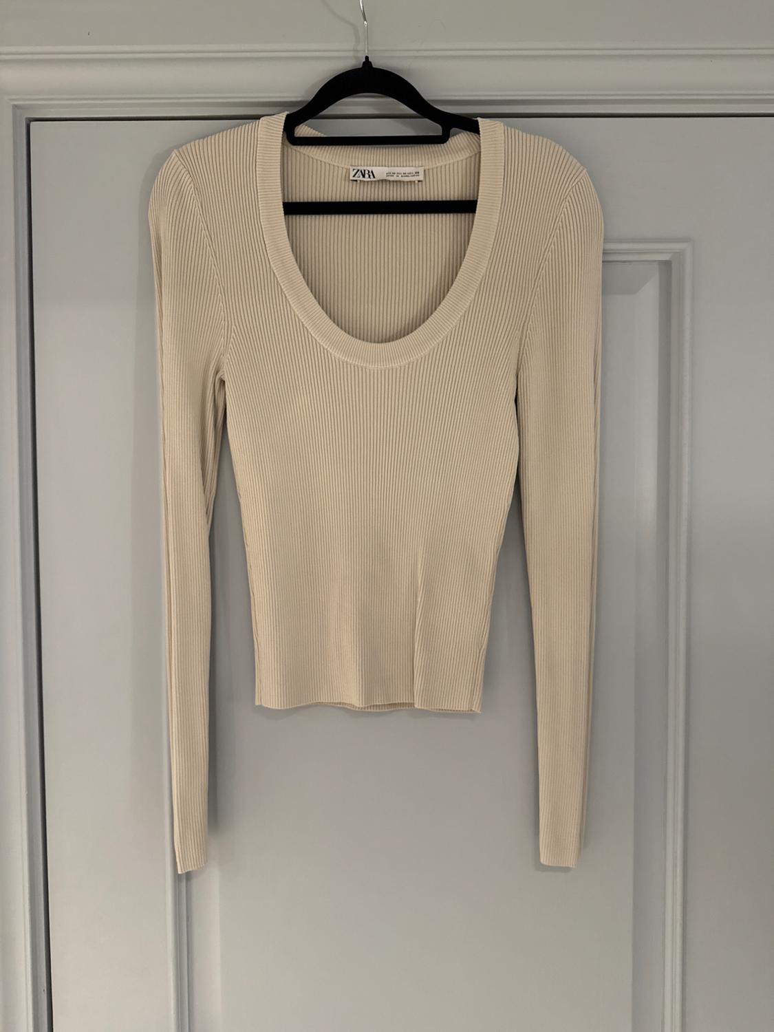 Beige knitted jumper with a round neck and long sleeves hanging on a black hanger over a grey door. Label visible.