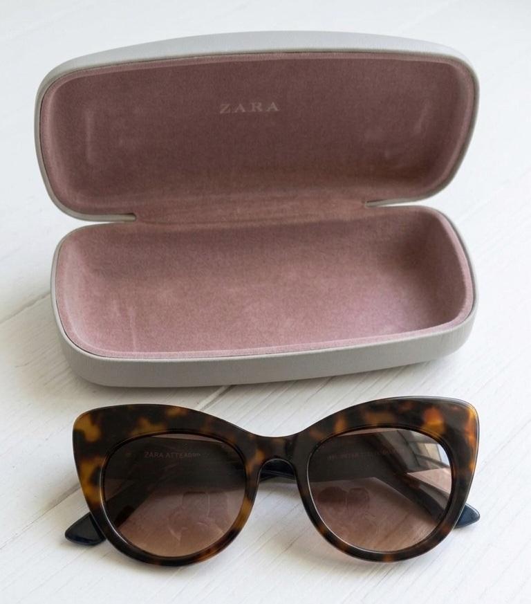 Tortoiseshell cat-eye sunglasses next to an open grey case with a pink interior and the text ZARA, on white wood.