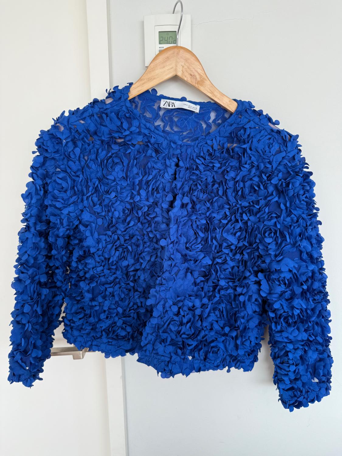 Blue jacket with a floral texture hanging on a wooden hanger. Visible label with the brand "ZARA". Minimalist white background.