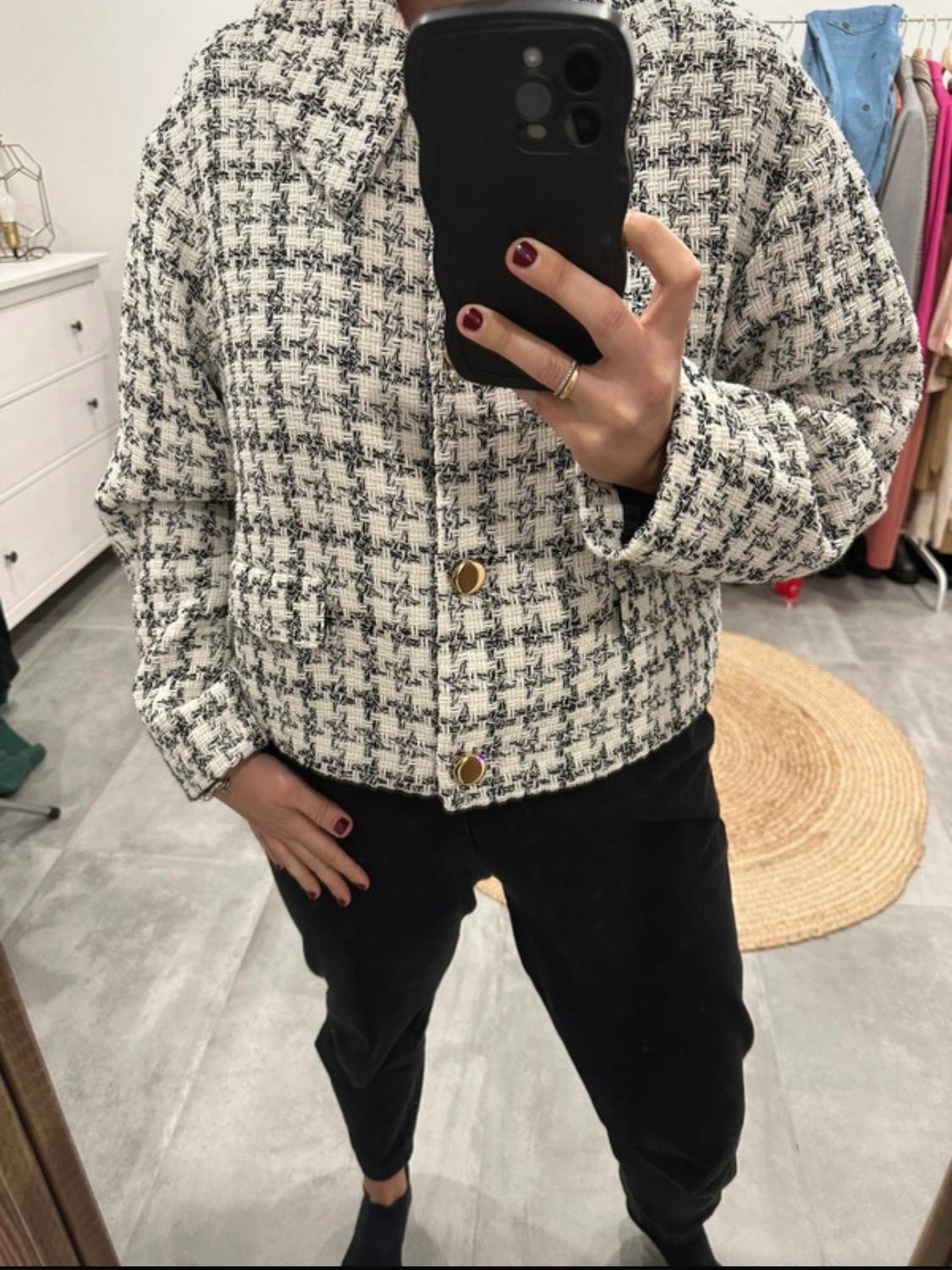 Mirror selfie with a black mobile phone. Person wearing a black and white textured checked jacket with gold buttons and black trousers in a dressing room.