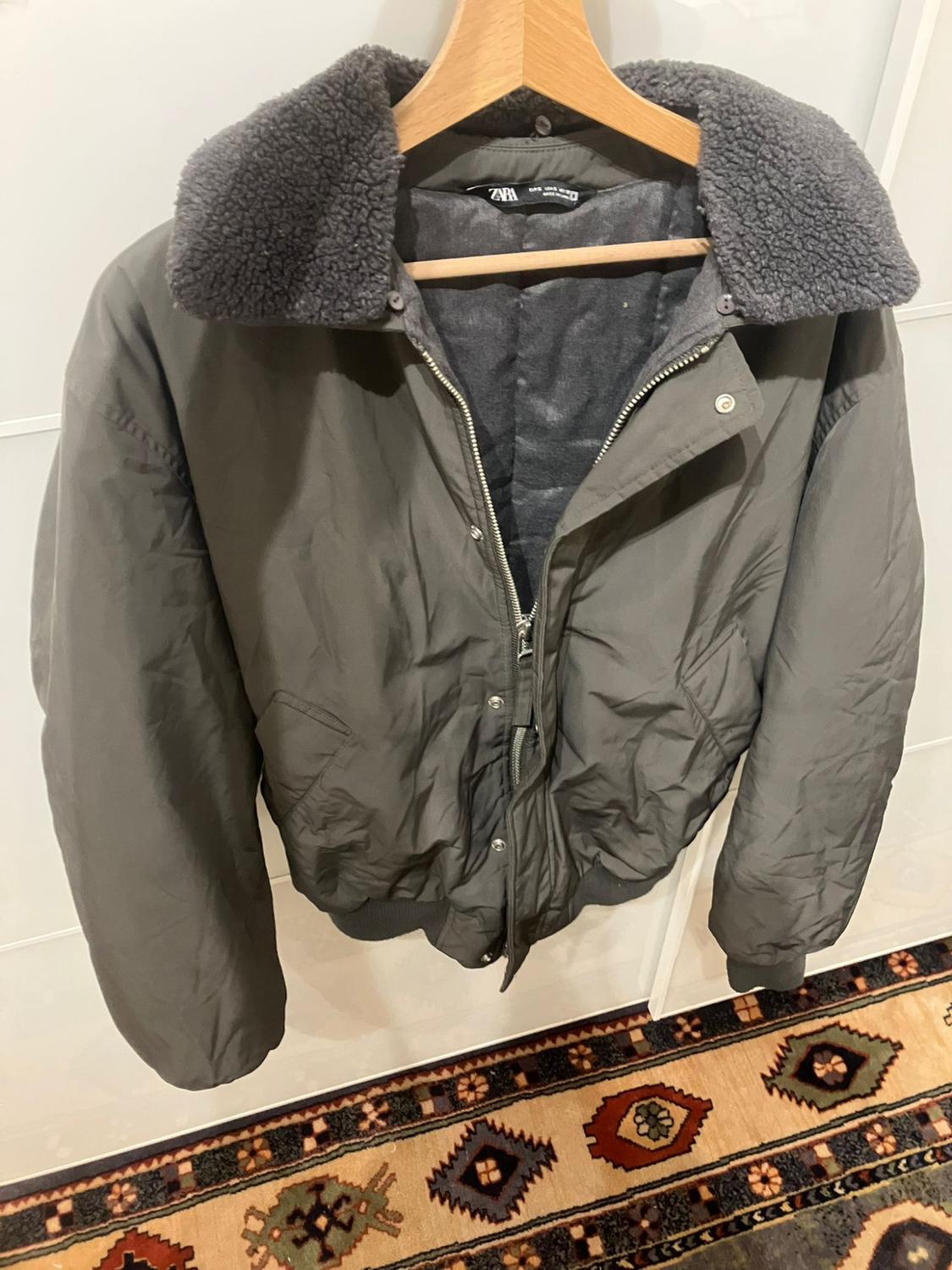 Grey padded jacket with a wool collar, hanging on a wooden hanger. Background with a geometric design rug.