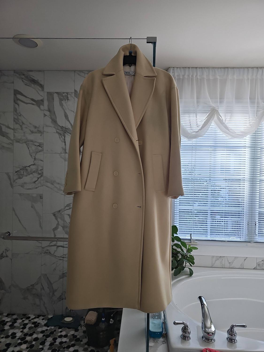 Beige double-breasted long coat hanging in a bathroom with a bathtub, marble wall, and window. ZARA text on the label.