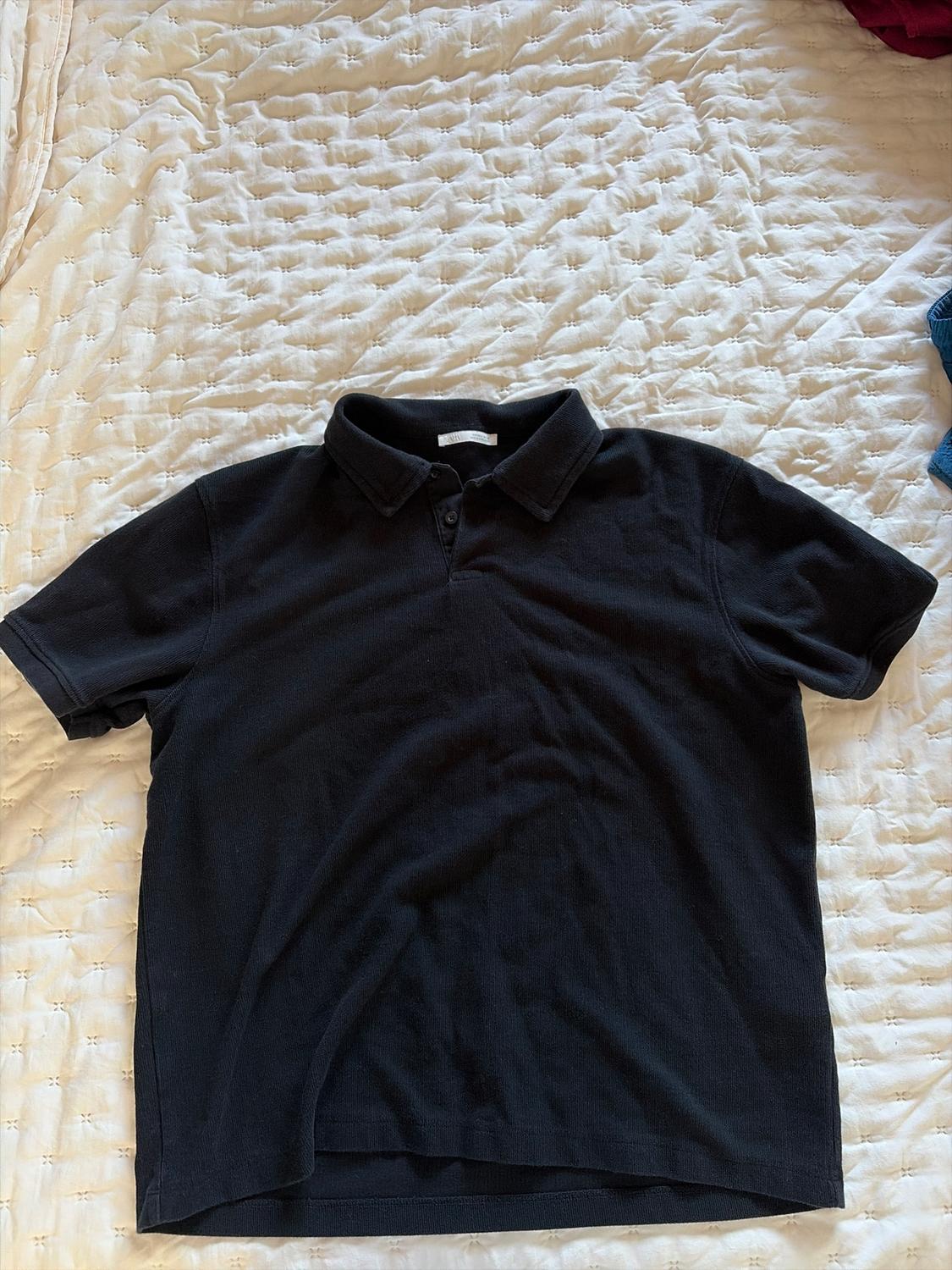 Black short-sleeved polo shirt on a white quilted bedspread. The polo shirt has a classic collar and two visible buttons.