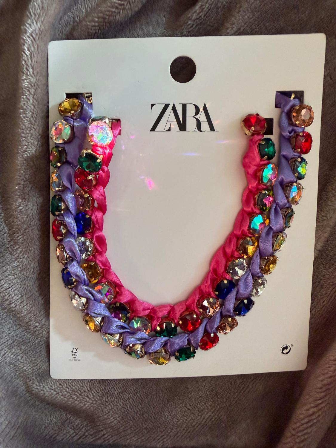 ZARA necklace made of pink and purple braided ribbons with multicolor rhinestones, on its cardboard tag over a soft, textured blanket.