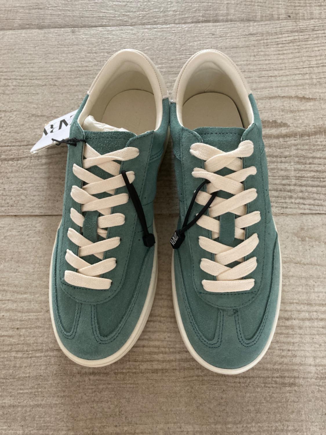 Mint green trainers with white laces on light wood flooring. Label visible on the left shoe.
