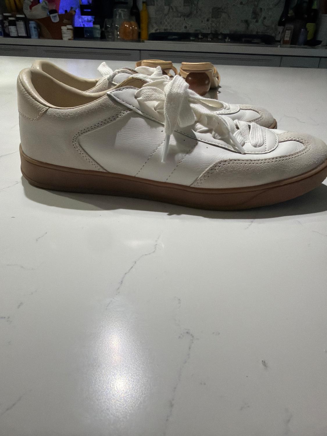 White sneakers with suede details and a brown sole on a white marble countertop, in a relaxed setting with a kitchen background.