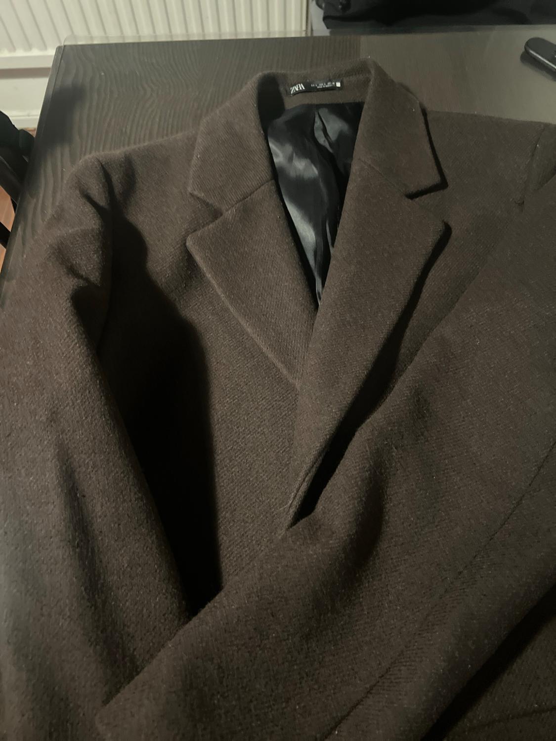 Dark brown coat with lapels and a black inner lining, resting on a table. The neck label reads ZARA.