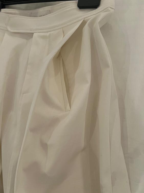 White pleated trousers hanging on a black hanger, showing fabric details and design against a light background.