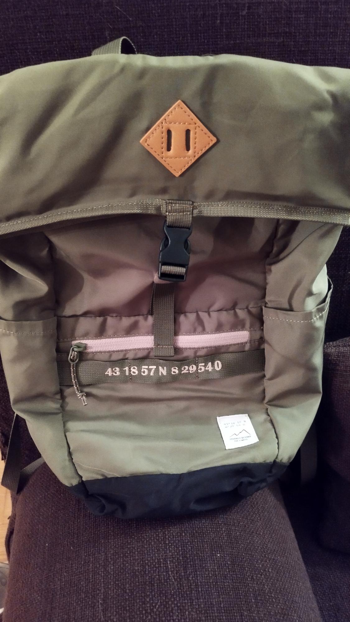 Olive green rucksack with black zip and brown patch. Visible text: "43 18 57 N 8 29 540". Dark background.