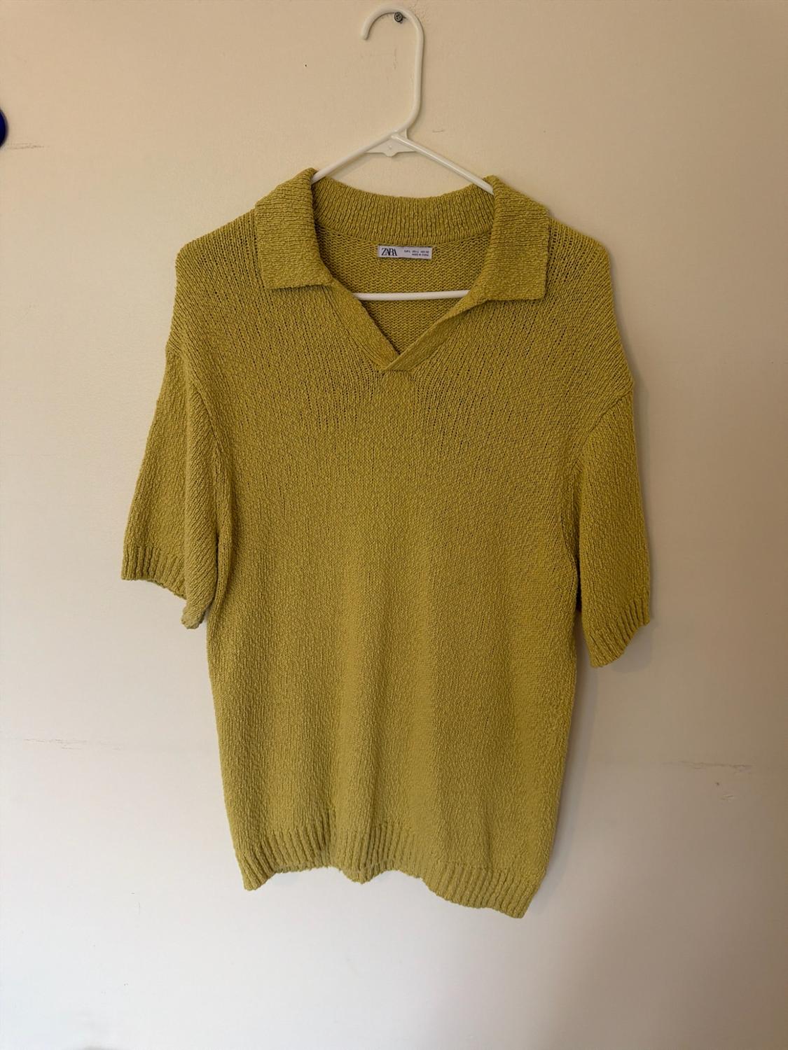 Mustard-coloured knitted polo shirt, short sleeves and V-neck. Hanging on a white hanger against a light-coloured wall. Label with the text Zara.