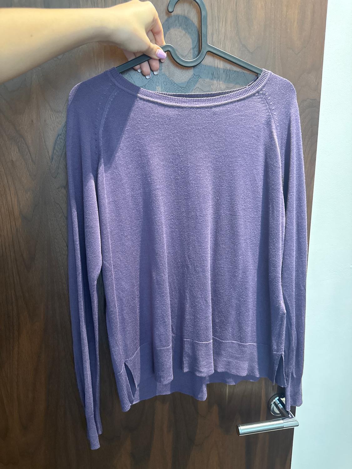 A purple long-sleeved jumper is hanging on a black hanger, held by a hand, in front of a wooden door.