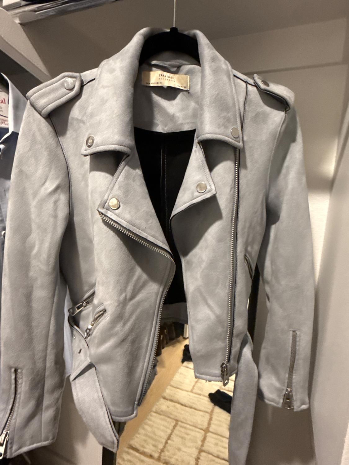 Light gray biker jacket with silver zippers and a belt, hanging in a closet. Interior label with the text ZARA BASIC.