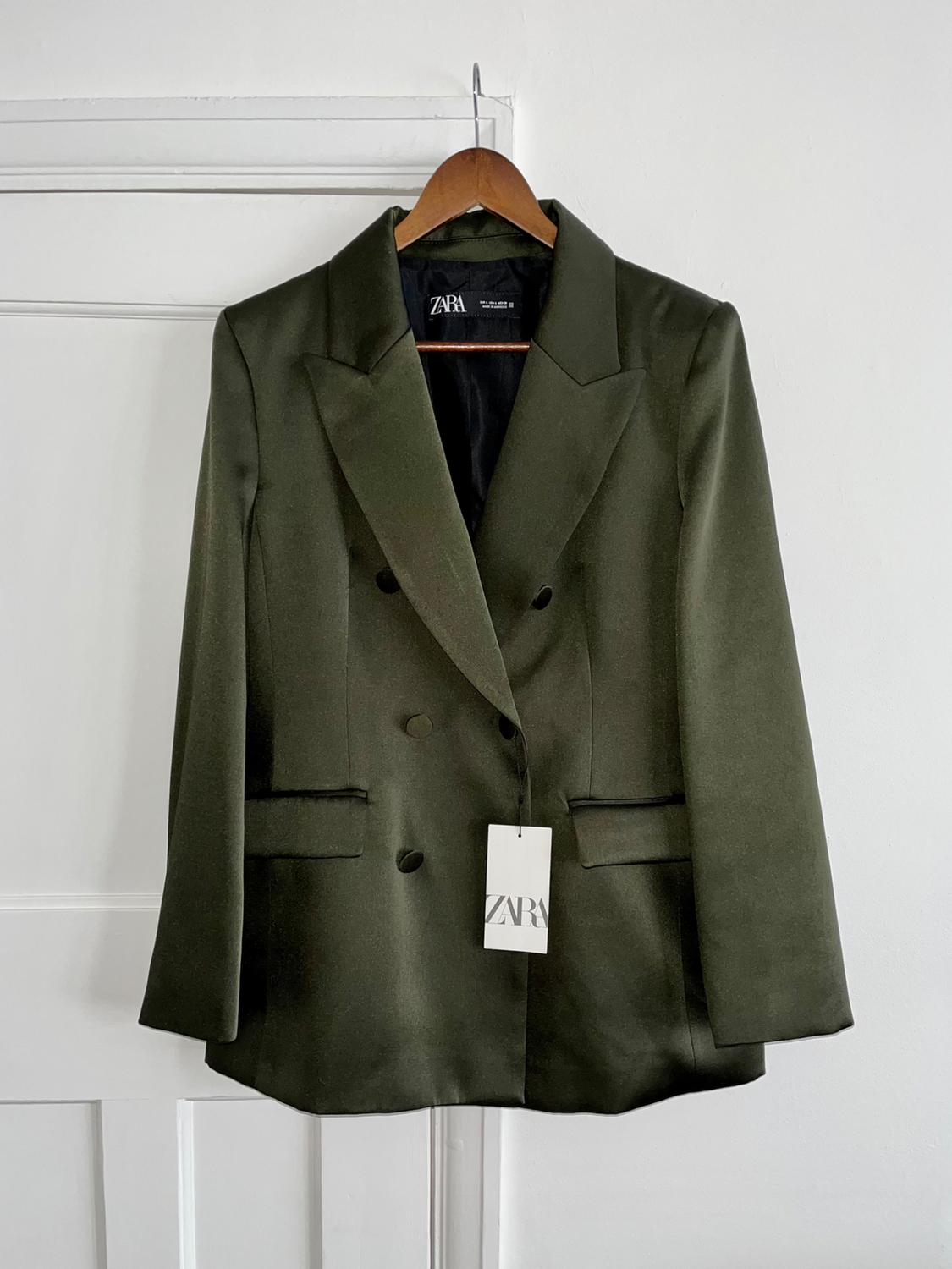 Olive green double-breasted jacket hanging on a wooden hanger. Visible label with the text "ZARA". White background.
