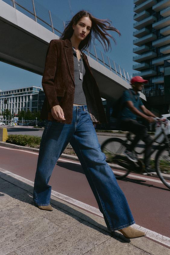 Hnědý blejzr s klopami a předními kapsami, šedé tričko a modré džíny širokého střihu.