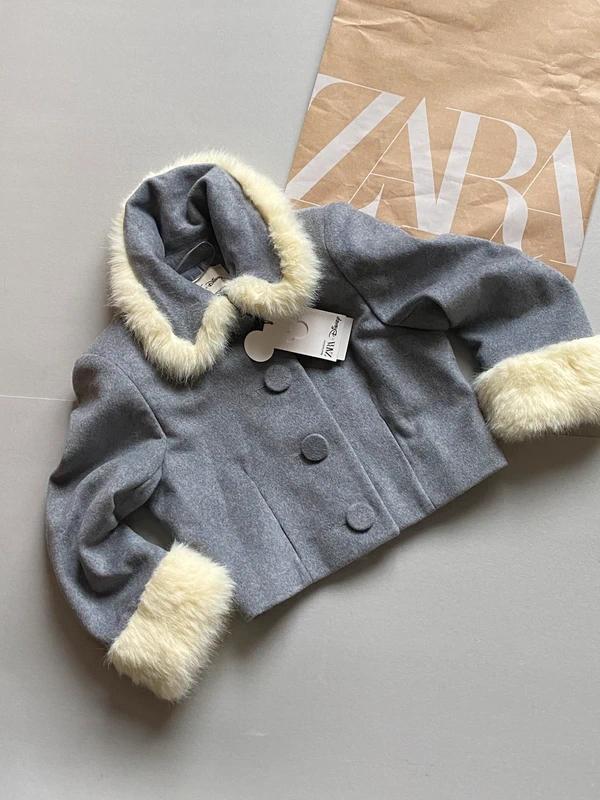 Grey jacket with a collar and white fur cuffs on a grey background. Visible label. Paper bag with "ZARA" text in the background.