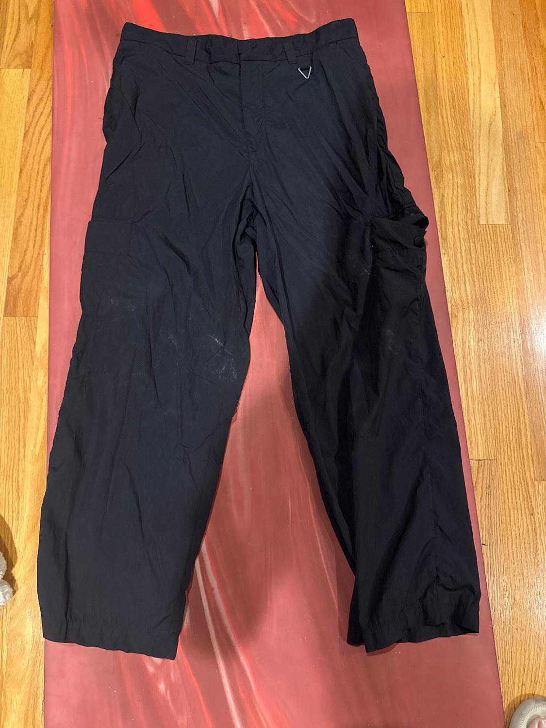 Black cargo trousers on a wooden surface and red carpet, with a small metal triangle on the waistband.