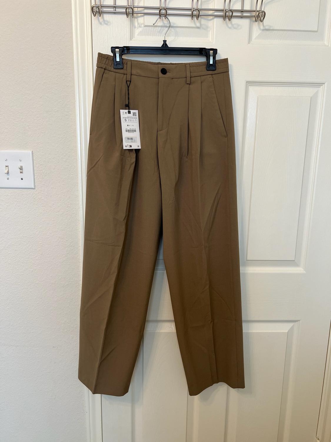 Brown pleated trousers hanging on a hanger over a white door. They have a label bearing the text S and 59.90.