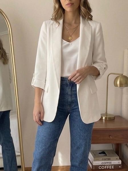 Woman wearing a white blazer, top and blue jeans. Relaxed setting with a mirror, gold lamp and books bearing the text COFFEE.