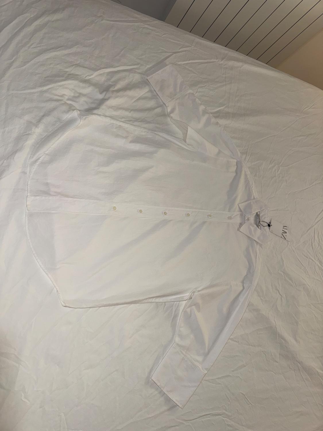 White long-sleeved button-up shirt, laid out on a bed with white sheets. Label visible on the collar.