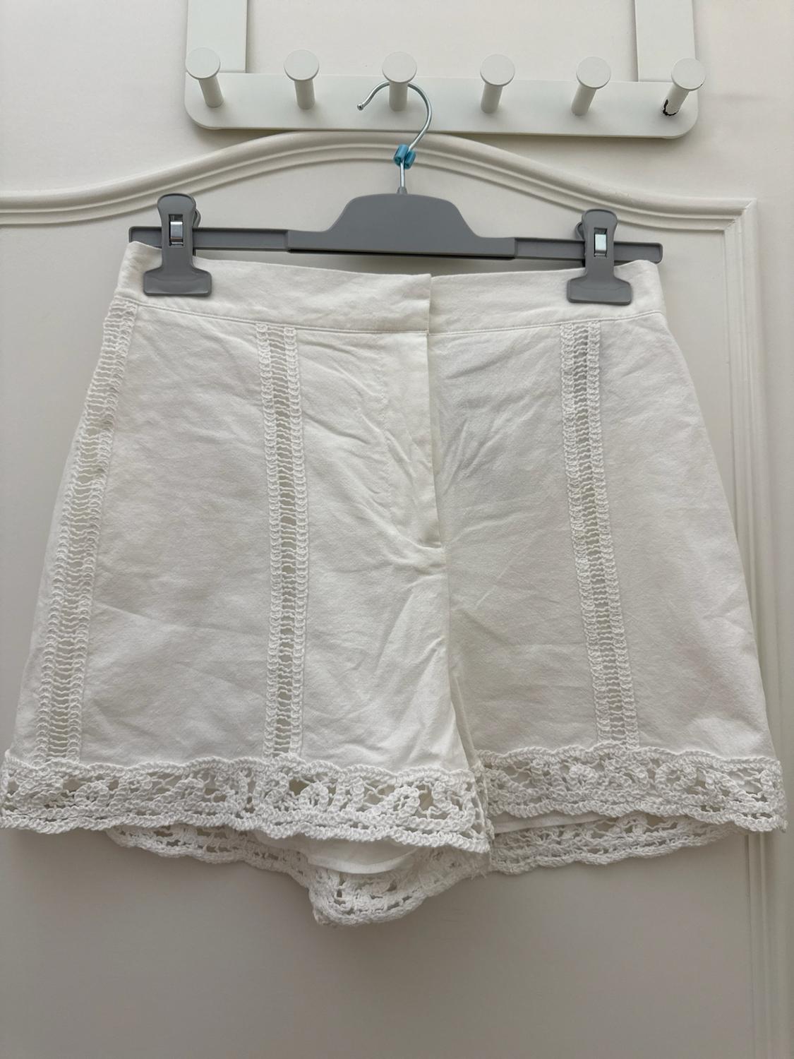 White shorts with vertical lace detailing and lace trim on the hem, hanging on a grey hanger in front of a white door.