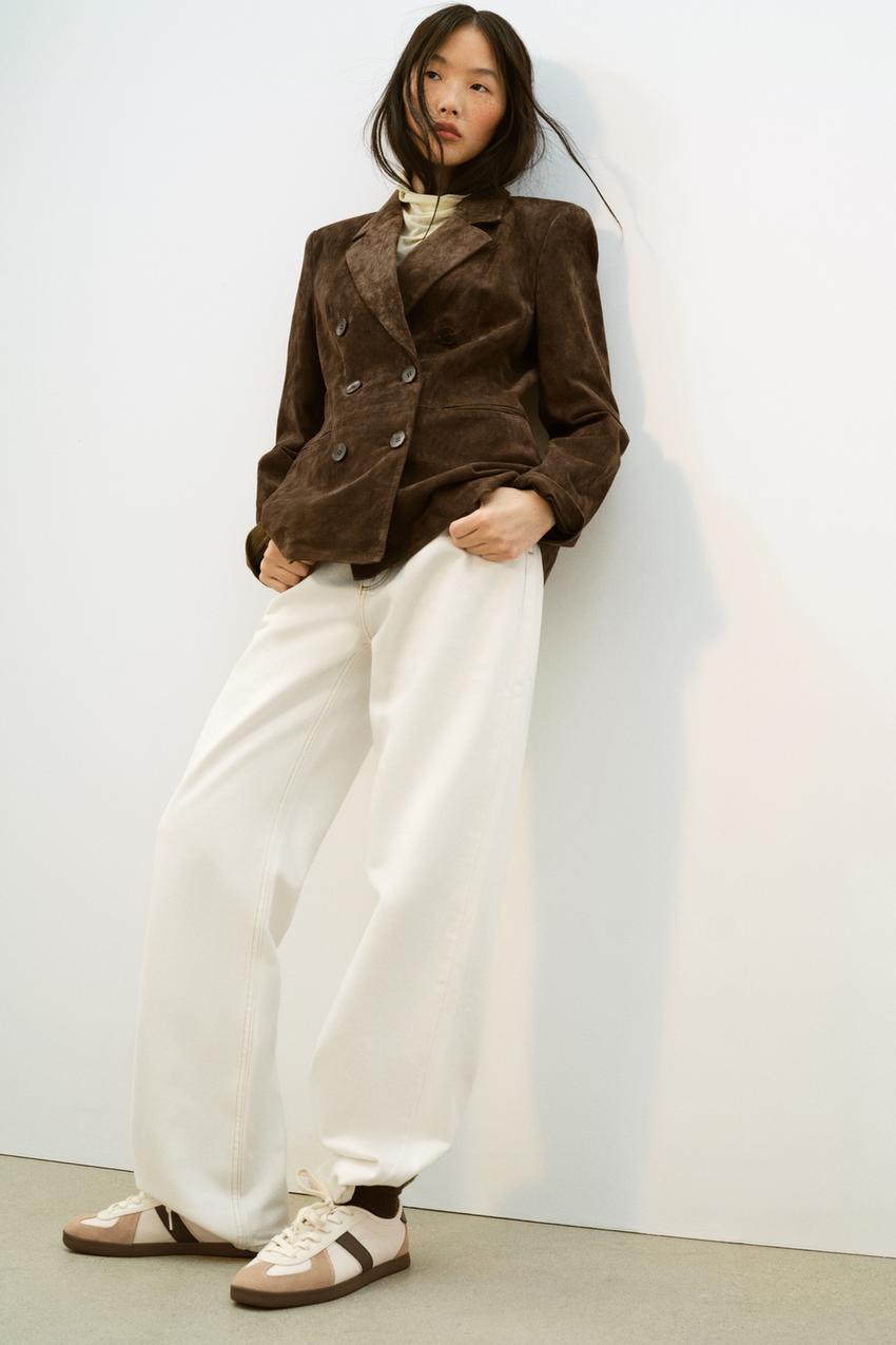 Conjunto de chaqueta marrón doble botonadura, pantalones blancos anchos y zapatillas deportivas marrones y blancas.