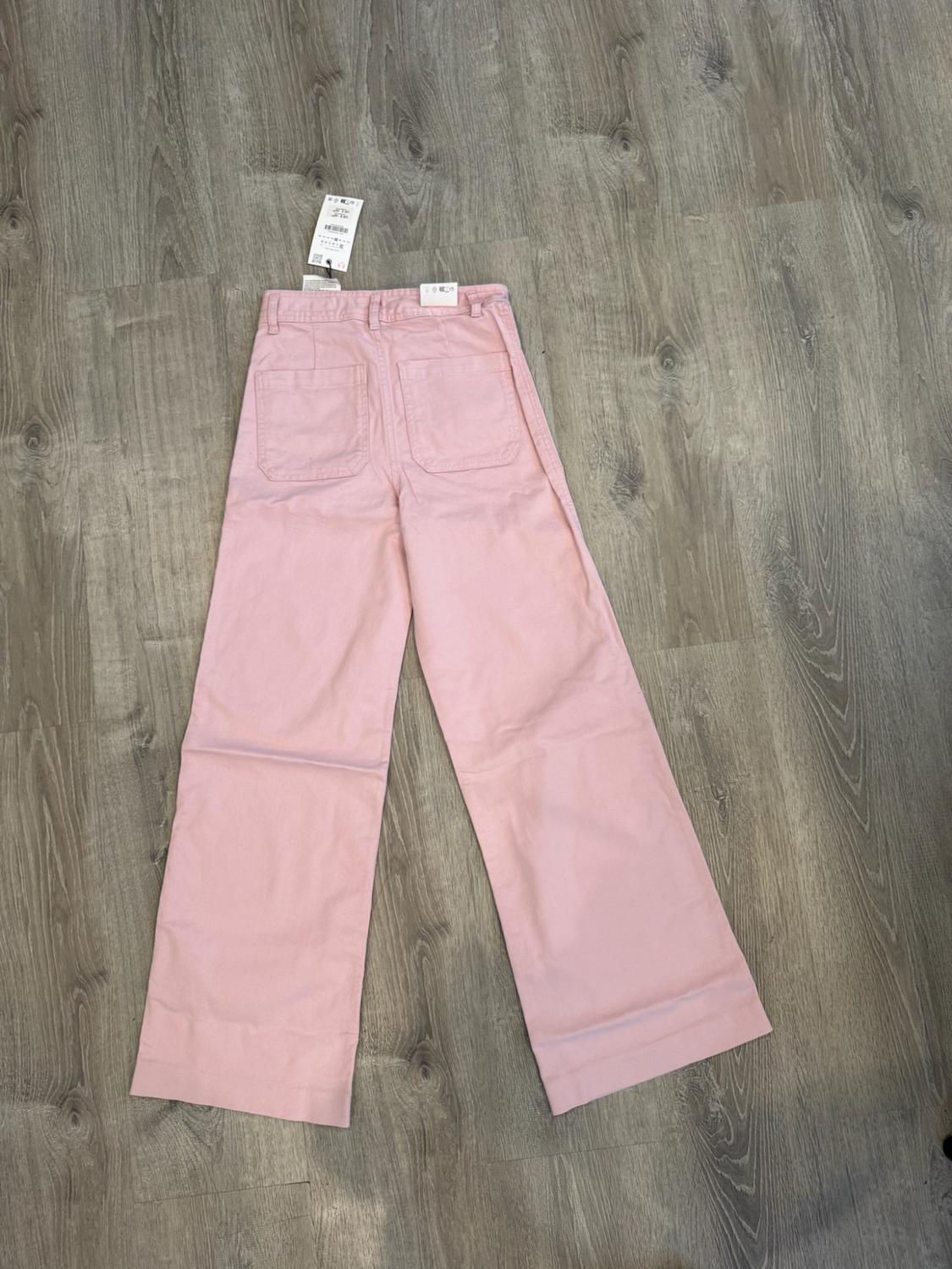 Light pink wide-leg trousers seen from the back, with patch pockets and labels, laid out on a wooden floor.