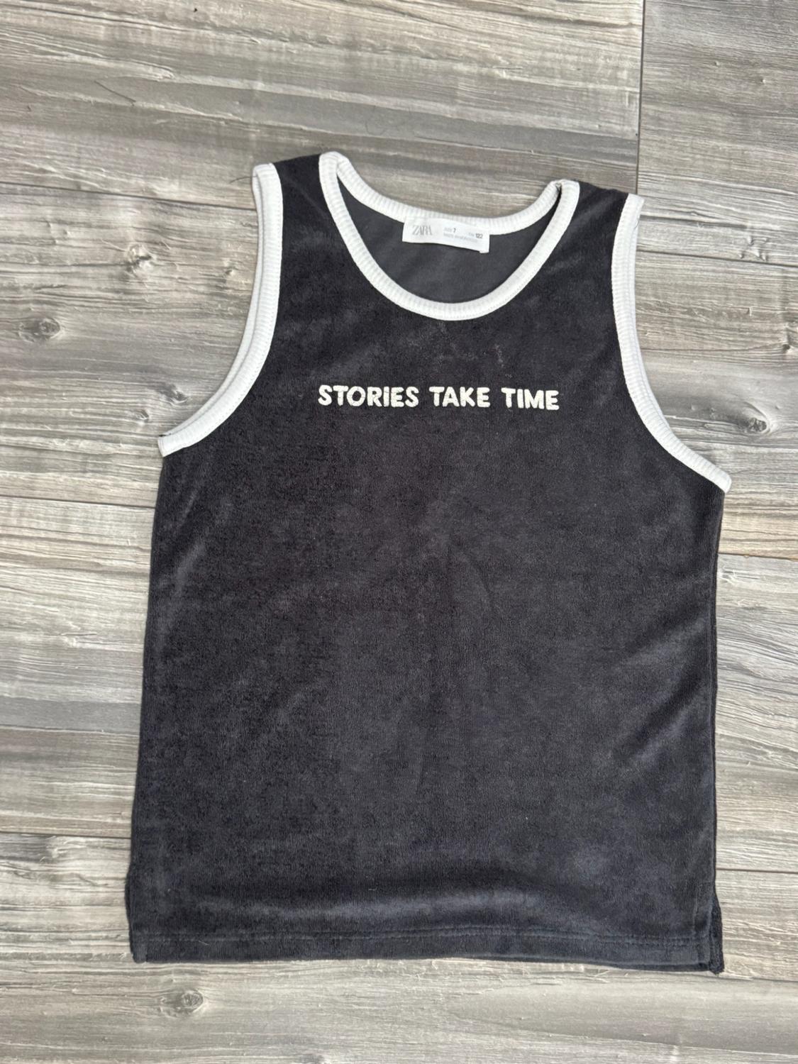 Black sleeveless T-shirt with white trims against a wooden background. Text: ZARA and STORIES TAKE TIME.