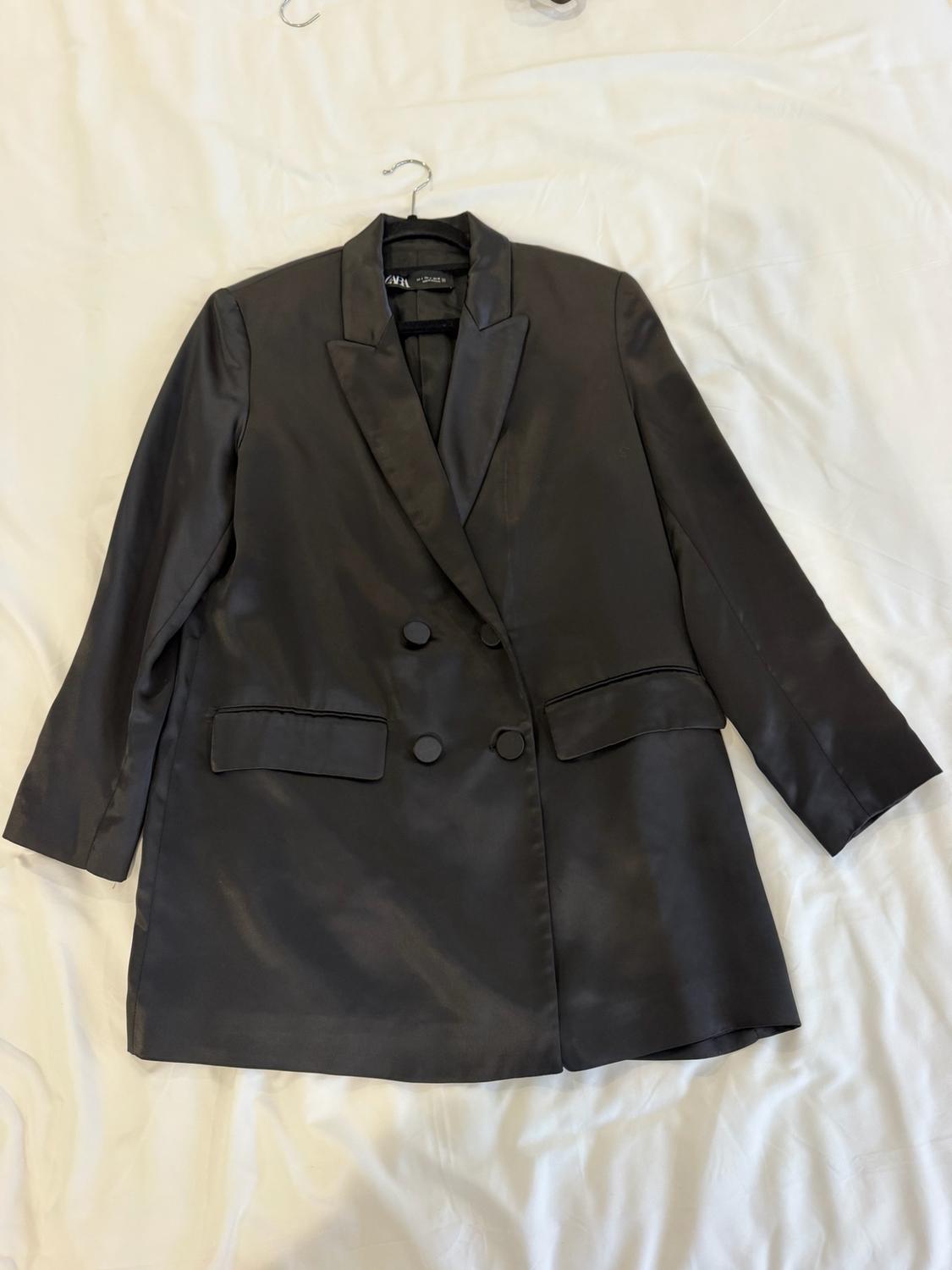 Black satin double-breasted blazer with buttons and flap pockets, on a wrinkled white sheet. ZARA label visible on the inside.