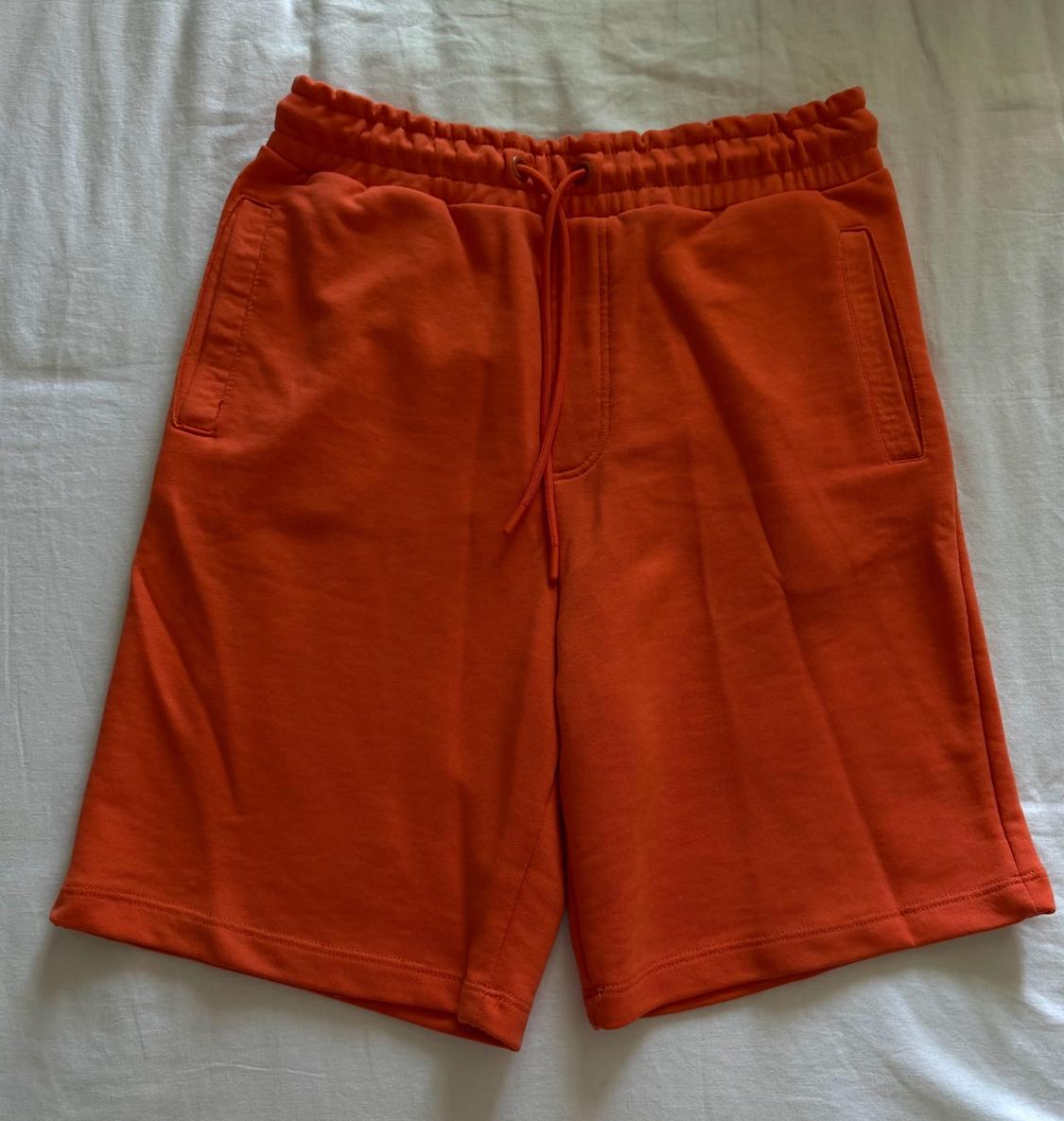 Orange sports shorts with an elasticated waistband, drawstring and side pockets, on a crumpled white fabric. Casual style.