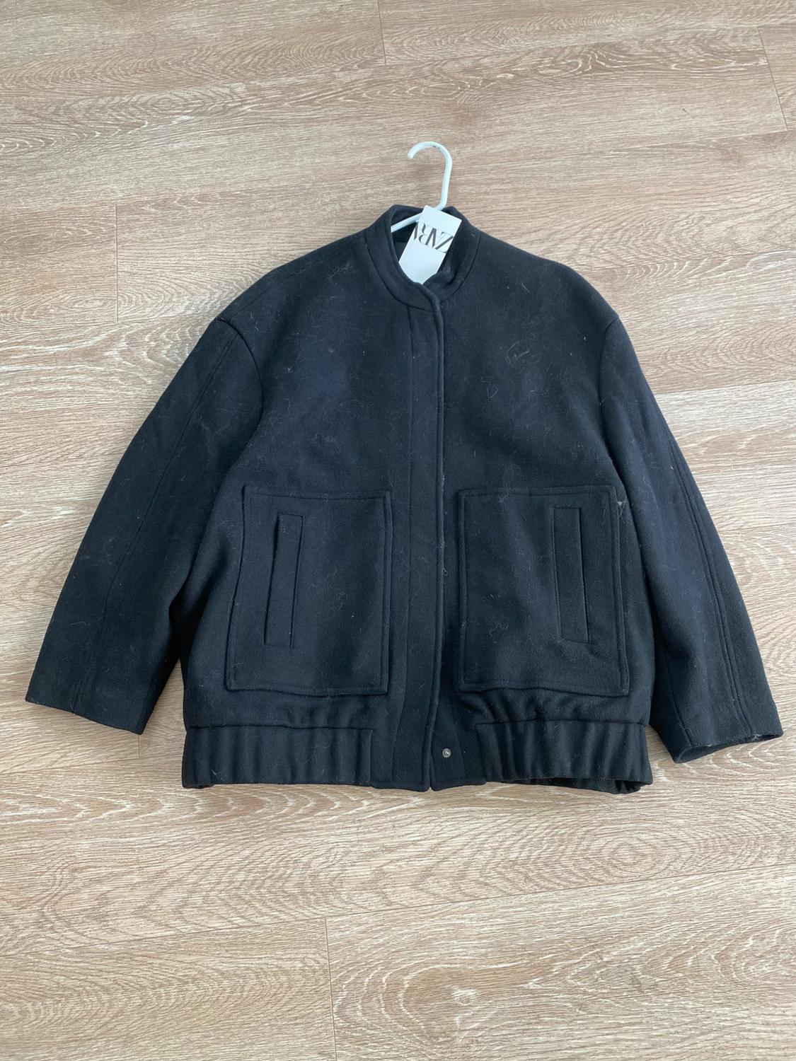 Black wool jacket with front pockets, hanging on a hanger over a light wood floor. Visible tag with "ZARA" text.