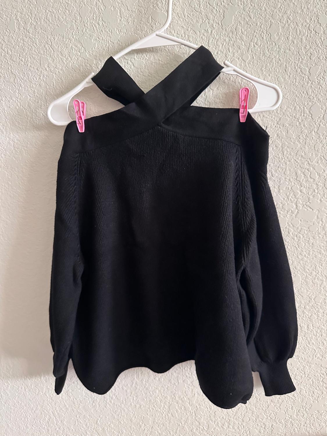 Black jumper with a crossover neck hanging on a white coat hanger, fastened with pink clothes pegs, against a light-coloured wall.