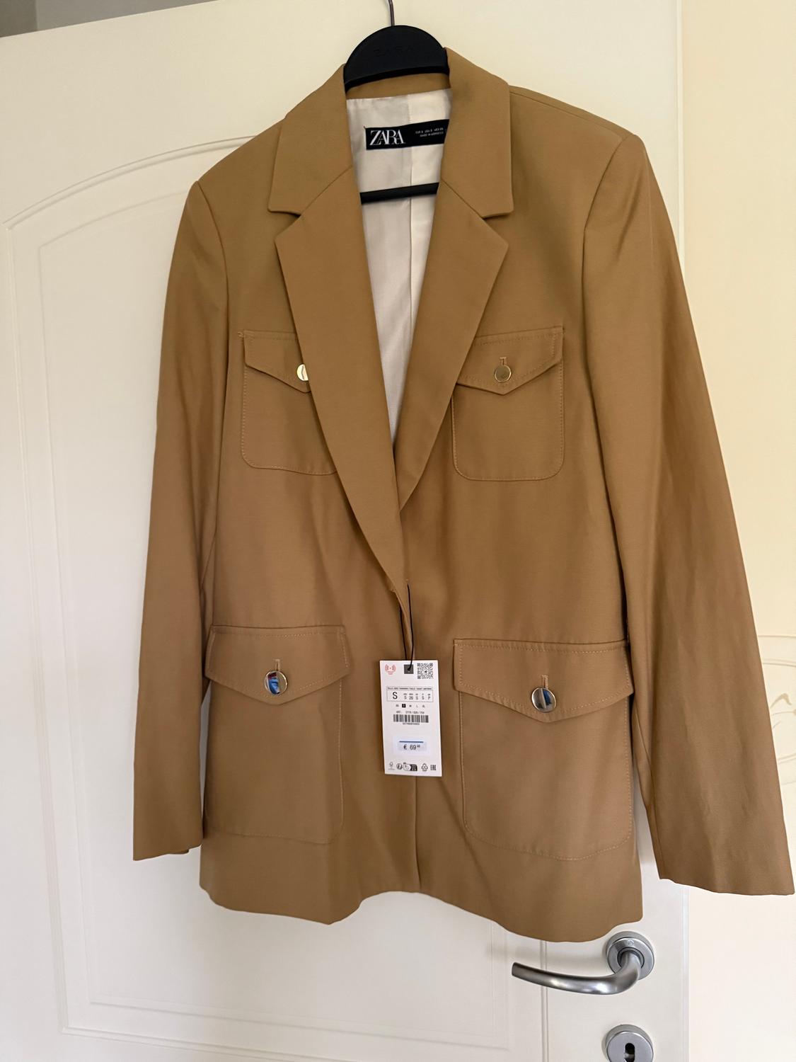 Beige Zara jacket hanging on a white door, with front pockets and a visible label showing a barcode and size S.