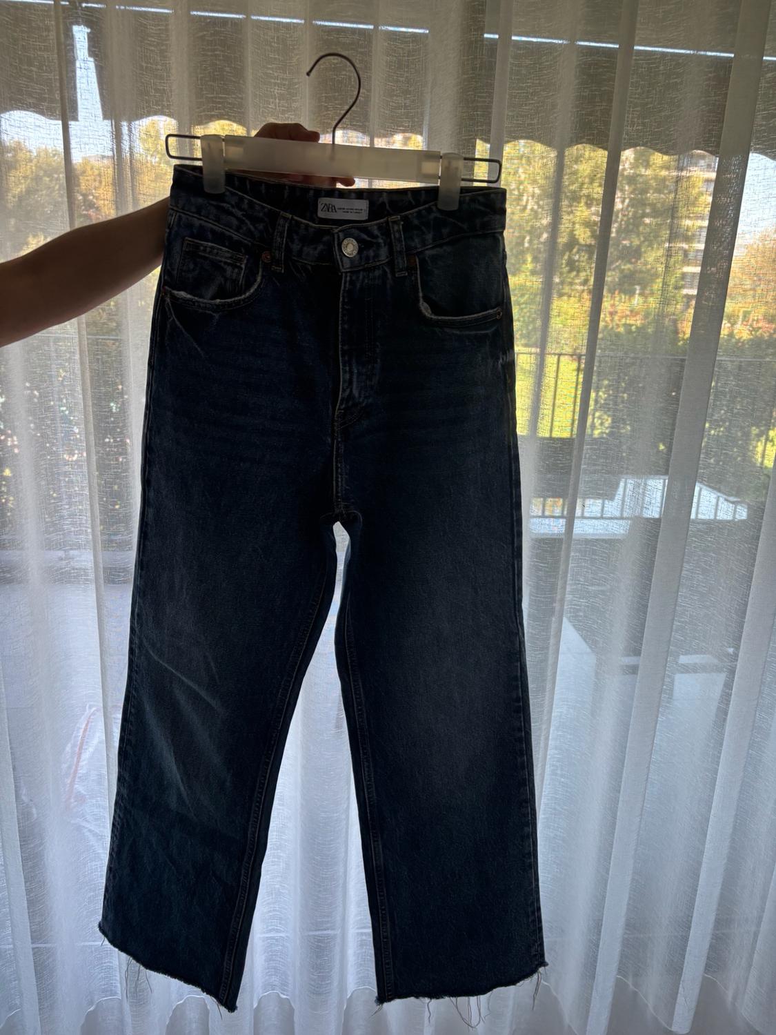 A hand holds a pair of dark blue jeans with a frayed hem on a hanger in front of a window with white curtains. ZARA label.